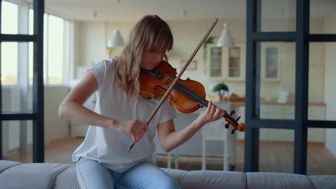 Teenage girl playing violin in living room. Female violinist playing chords on musical instrument at home. Inspired musician pressing strings of violin. Young woman sitting on sofa