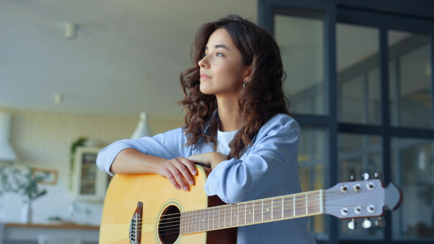 Dreamy girl holding acoustic guitar at home. Portrait of happy woman looking at camera. Smiling lady dreaming in living room. Female musician posing at camera with string instrument