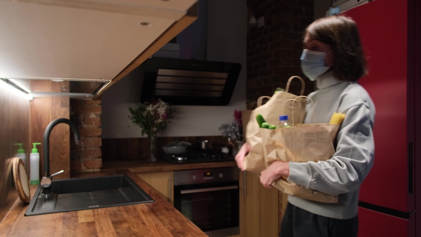 Young short haired housewife puts large paper bags with products on countertop and takes off protective mask in kitchen at home