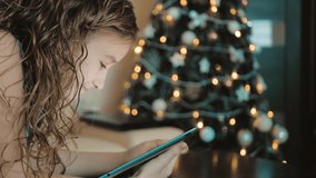 Adorable little girl plays on a tablet, at background the christmas tree illuminated with christmas.