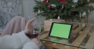 Young caucasian woman wrapping presents gift box with ribbon on table at home. Girl enjoying wine chatting with friends or family on tablet with green screen