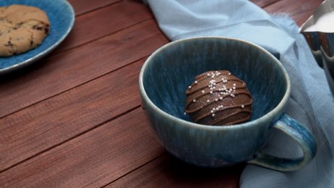 Cocoa bomb that melts with hot milk.   Milk splash in slow motion. Cup of dark hot chocolate. Comfort food. Wood table with blue cloth and a cookie. 