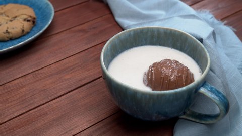 Cocoa bomb that melts with hot milk.  Opens in slow motion and marshmallows pop. Cup of dark hot chocolate. Comfort food. Wood table with blue cloth and a cookie. 