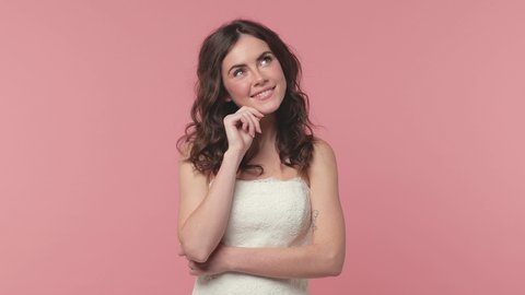 Smiling pensive bride young brunette woman 20s years old in beautiful white wedding dress posing isolated on pink pastel background studio. Wedding concept. Put hand prop up on chin looking aside up