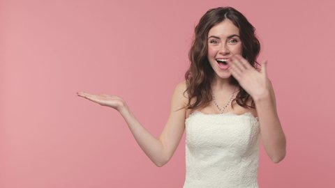 Surprised bride young brunette woman 20s in beautiful white wedding dress posing isolated on pink background studio. Wedding concept. Pointing index finger on empty palm say wow cover mouth with hand