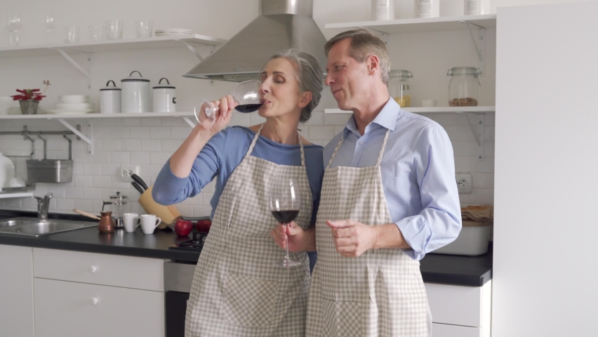 Happy affectionate older middle aged family couple wearing aprons having fun, dancing in kitchen. Senior husband and wife drinking wine enjoying dance celebrating Valentines day or anniversary at home Royalty-Free Stock Footage #1065783715