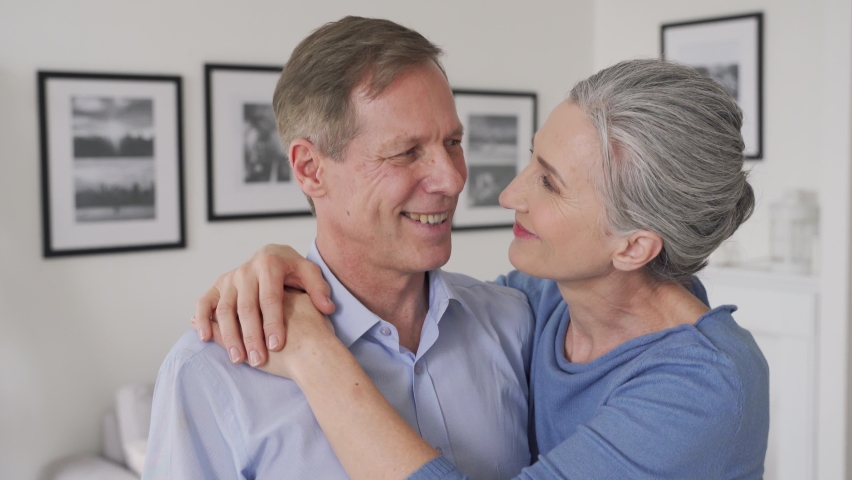 Happy senior mature 60s affectionate family couple hugging looking at camera, standing in apartment. Smiling mid aged husband and wife embracing posing for portrait at home enjoying love and wellbeing Royalty-Free Stock Footage #1065783721