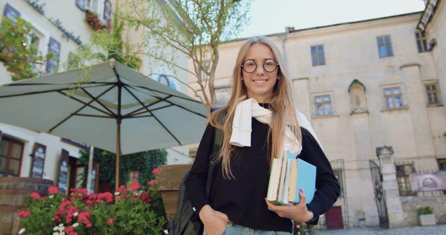 Attractive cheerful modern female student with books and backpack posing on camera near beautiful street cafe on the road to university