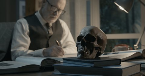 Academic professor sitting at office desk, he is studying a human skull and writing a report