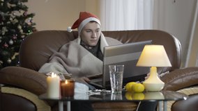 Portrait of ill Caucasian young man using video chat on laptop on Covid-19 New Year. Sick handsome guy celebrating Christmas eve alone at home sitting on couch. Coronavirus quarantine concept.