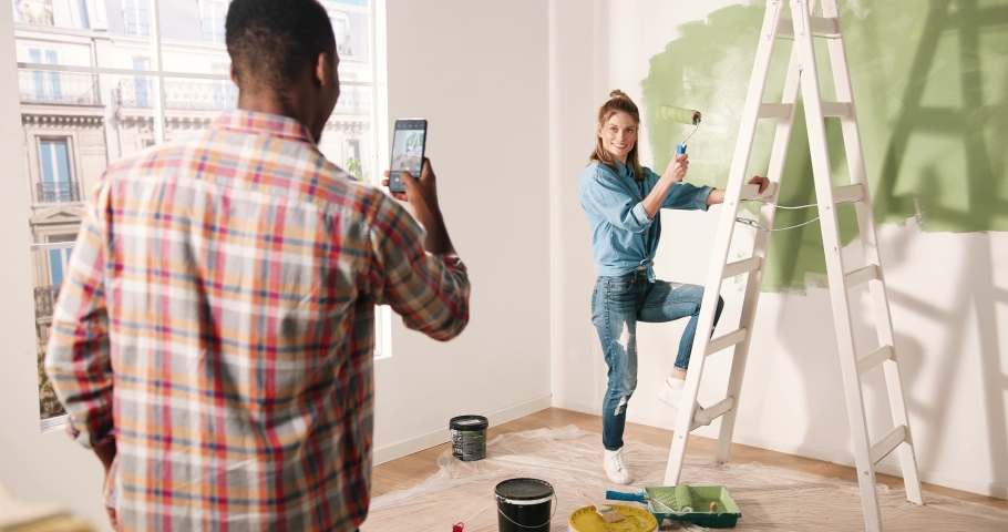 Happy multinational couple making repairs in new apartment. Husband taking photo of his wife on smartphone. Cheerful woman posing with paint roller near ladder.