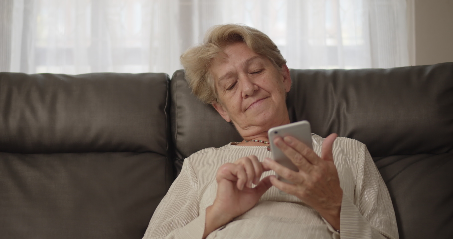 An elderly woman after retirement cut short hair with light brown hair Lie on the sofa laugh and enjoy social media on her smartphone restlessly at her home alone. Royalty-Free Stock Footage #1065812989