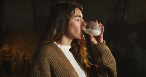 Cinematic close up shot of young female farmer drinking fresh just produced biological milk used for genuine dairy products industry and smiling satisfied in cowshed stable of countryside dairy farm.