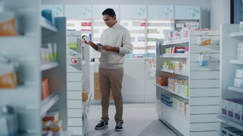 Pharmacy Drugstore: Portrait of Handsome Young Indian Man Using Smartphone, Searching to Purchase Best Medicine, Drugs, Vitamins. Shelves full of Health Care, Wellness, Sports Supplements