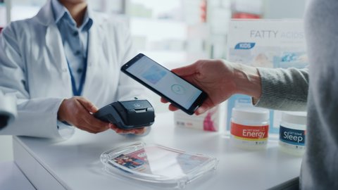 Pharmacy Drugstore Checkout Cashier Counter: Pharmacist and a Customer Using NFC Smartphone with Contactless Payment Terminal to Buy Prescription Medicine, Health Care Goods. Hand Focus Closeup
