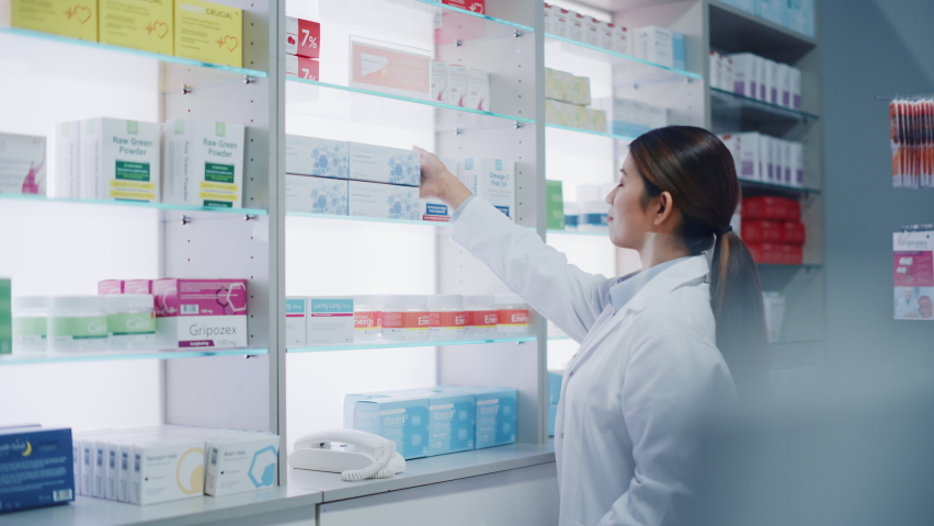 Pharmacy Drugstore: Portrait of Beautiful Diligent Asian Pharmacist Wearing White Coat Arranges Medicine Packages, Drug Boxes, Vitamin and Supplement Pills on a Shelf Behind the Counter | Shutterstock HD Video #1065814336