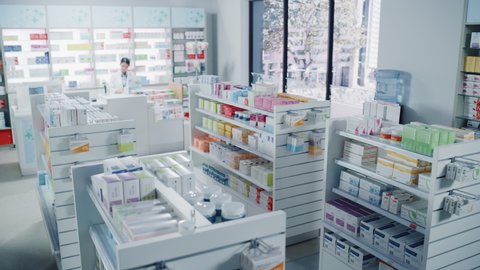 Big Modern Pharmacy Drugstore with Shelves full of Packages Full of Modern Medicine, Drugs, Vitamin Boxes, Pills, Supplements, Health Care Products. Pharmacist Standing at Counter. Establishing Shot