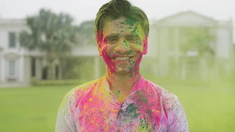 Close front view  movement shot of powdered colors blown in the air falling over a young handsome Indian male smiling and looking at the camera while celebrating Holi festival. 