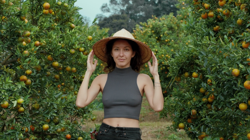 girl standing and smiling in an orange orchard wearing a vietnamese hat looking straight at the camera. cinematic shot bmpcc 6k