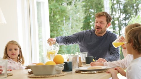 Father pouring orange juice as family wearing pyjamas sit around table enjoying pancake breakfast together - shot in slow motion