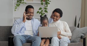 Home portrait of young African American family parents with small daughter sitting in living room interior in cozy house looking at laptop screen watching movies cartoons video online through computer