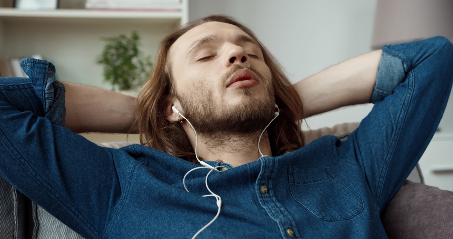 Young handsome caucasian guy with long hair in denim shirt is resting lying on couch and listening music on headphones.