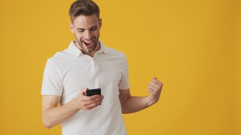 Handsome young excited man using mobile phone and making winner gesture isolated over yellow wall background