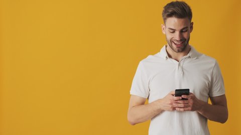 Handsome young surprised happy man using mobile phone isolated over yellow wall background