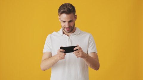 Handsome young cheerful man playing games by mobile phone isolated over yellow wall background