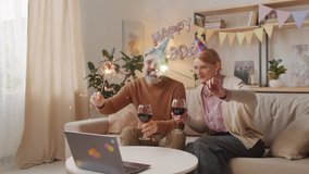 Medium shot of smiling and laughing middle-ages couple holding burning sparklers and toasting with glasses of wine while having online birthday party via laptop during quarantine