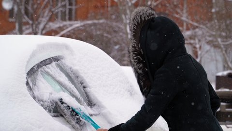 Woman in a Sheepskin Coat with a Hooded Fur Cleans the Car of Snow in Winter in Cold Weather. It is snowing. High quality 4k footage