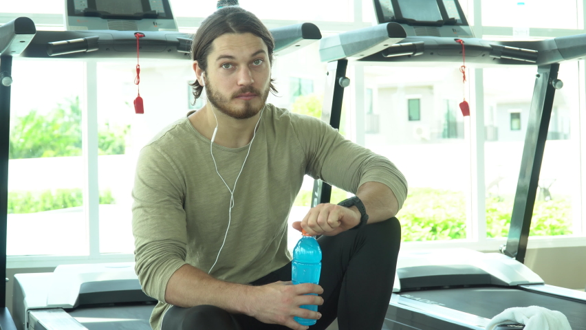 Attractive young sports man taking a break from running drinking Blue water energy drink on a treadmill in fitness gym indoors. runner male in sportswear workout or training on running machine