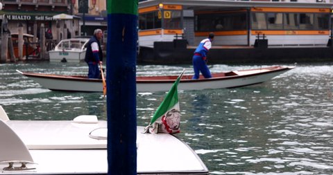 Italian flag attached to a rear of a motorboat with a water bus stop and two men steering a boat while standing on grand canal slow motion Venice Italy