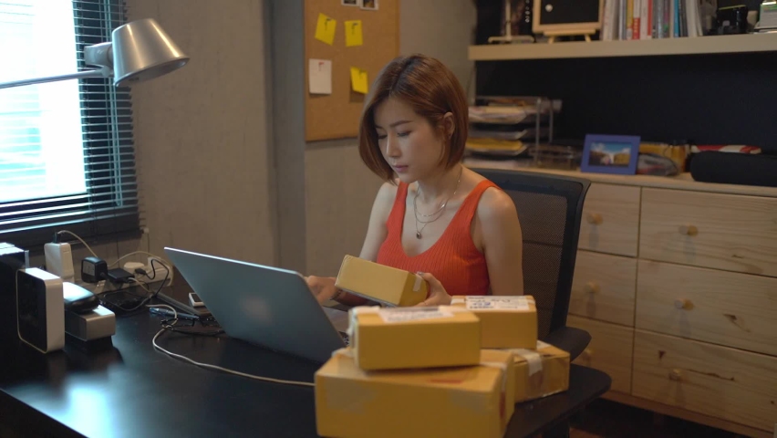Young short-haired Asian woman in orange tank top sitting at the desk checking order and address of each parcel box that will be shipping to the customer from online sales with laptop at a home office