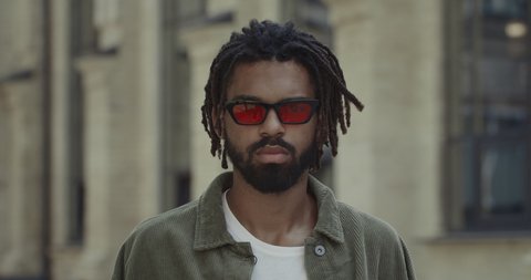 Crop view of young man with beard turning head and looking to camera. Portrait of afro american guy with dreadlocks wearing stylish glasses while standing at city street.