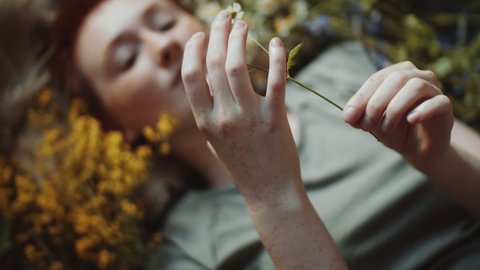 High angle rack focus shot of young dreamy woman with short red hair and freckles lying on burlap fabric among wildflowers and holding chamomile
