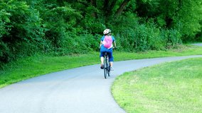 A cyclist rides a bike on a curved and paved bicycle path, lined by green trees and vegetation. Hand held clip from from behind rider.