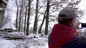 Speaking man in winter forest on top of a hill, while filming the small Swiss village Brugg at the foot of the hill. Real time panning shot of a middle aged man with red winter jacket and gray cap.