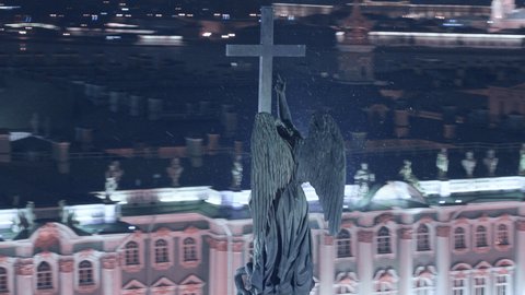 Aerial close view of the statue of an angel holding a cross on the Alexander Column in the city centre of St. Petersburg. Palace square, Winter Palace (Hermitage), panoramic view of the city at night.