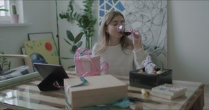 Young caucasian woman wrapping Christmas presents gift box with ribbon on table at home. Girl enjoying wine watching lessons on tablet or chatting with friends or family