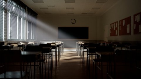 Rays of light falling to the empty dark classroom. The camera moves ahead passing by rows of desks and chairs. Teaching class without students during a break. Abandoned school. Disturbing mood.