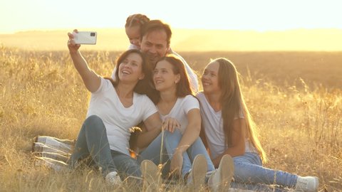 Mother and father with their daughters are photographed in field. Family of bloggers. Happy family travels. Selfie on smartphone in park. Mom, dad and children are sitting on a blanket filming a video