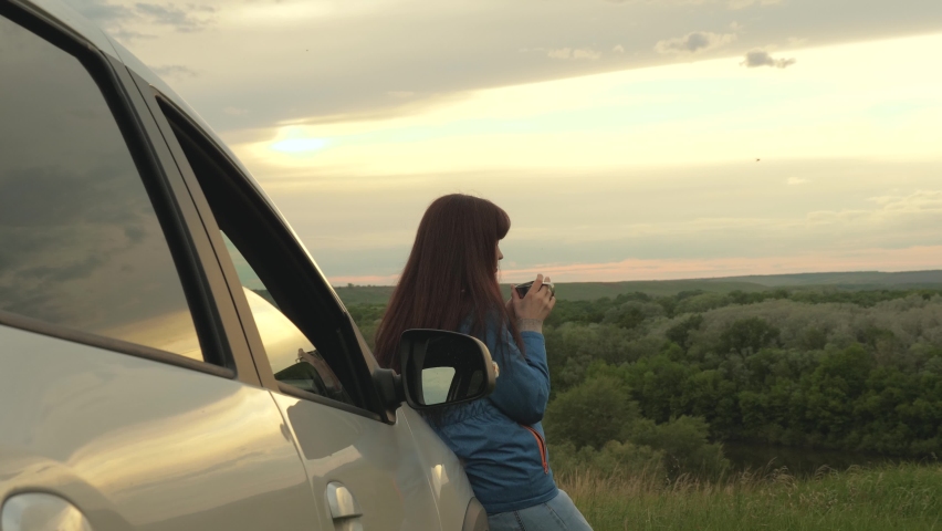 Woman driver traveler resting, drinking morning coffee from a mug in the sun. A tourist girl holds mug of hot tea in her hands and looks at sunset next to car. Freedom of travel and tourism by car. | Shutterstock HD Video #1065853990