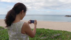 Asian woman on holiday vacation travel capturing beautiful pastel blue sea water n calm ocean wave crashing white sandy beach on cloudy sky by mobile cell phone or smartphone, 4k b-roll footage video