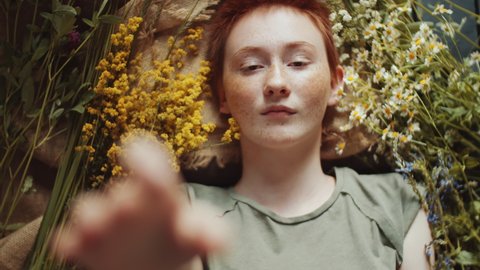 Top down shot of young beautiful woman with short red hair and freckles lying on burlap fabric among wildflowers and reaching out her arm towards the camera