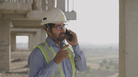 Shot of a young male Asian civil engineer in safety jacket and helmet is wearing eye glasses and talking on a mobile phone standing on top of under development building near construction site