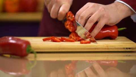  Man in kitchen is Slicing Red Paprika. Chef's hand in uniform cutting red fresh pepper  on glossy table. Close view. Healthy Food Concept. Preparing Vegetable for cooking. High quality 4k video.