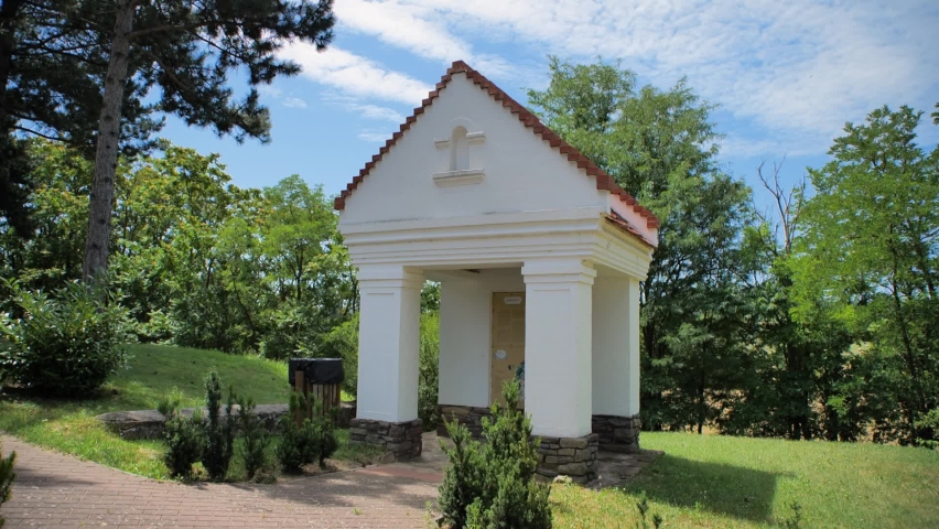 Small chapel in the garden of a church. Hungarian village grove and rural environment. Lush foliage and trees on a warm summer day. Kékkút, Hungary.