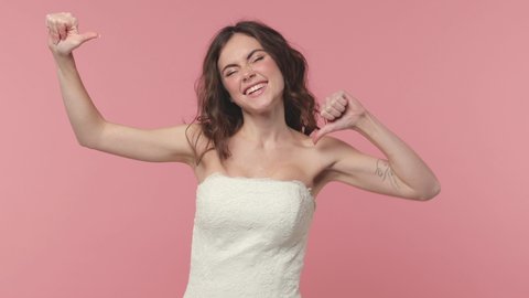 Excited cheerful bride young brunette woman 20s years old in beautiful white wedding dress posing isolated on pink pastel color background studio. Wedding concept. Pointing thumb on herself blinking