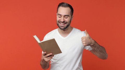 Smiling cheerful bearded young man 20s in white casual t-shirt isolated on orange color wall background studio. People sincere emotions lifestyle concept. Reading book showing thumb up like gesture